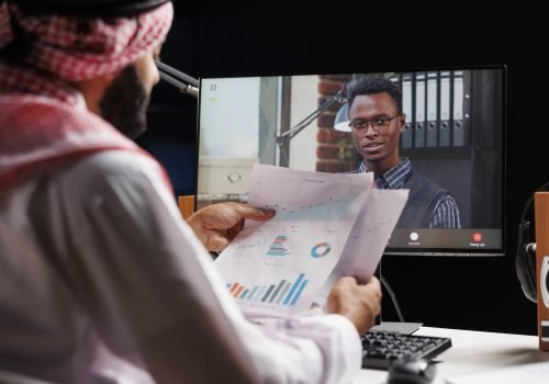 Computer screen takes center stage displaying video call between Muslim businessperson and African-American associate. Picture illustrates the utilization of modern technology by the two individuals.