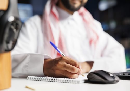 Close-up shot of a notebook and pen being used by a Middle Eastern man in traditional clothing seated at a table. Selective focus on Muslim guy writing down research notes.